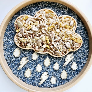 May include: A wooden tray with a blue and white speckled surface. A cloud-shaped container filled with pasta, beans, and small stones is in the centre of the tray.  There are white lightning bolt shapes and raindrops around the container.