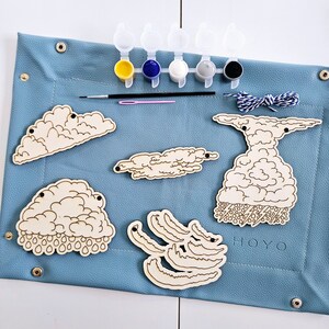 May include: A wooden craft kit with five paint pots, a paintbrush, a needle, and thread. The kit includes six wooden shapes of clouds, one with rain, one with lightning, and one with a fluffy texture. The kit is on a blue surface and includes the word "HOYO".