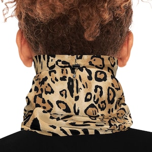 May include: A tan and black leopard print neck gaiter worn around the neck. The neck gaiter is made of a soft, flexible material. The person is wearing a black shirt. The background is white.