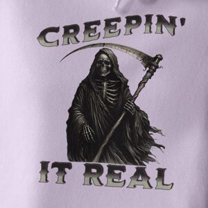 May include: A light purple hoodie featuring a graphic of a grim reaper holding a scythe. The text CREEPIN' is arched above the figure, and the text IT REAL is positioned below the figure in a dark, stylized font with a subtle gradient effect.