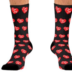 May include: A pair of black crew socks featuring an all-over repeating pattern of red hearts. Each heart contains the white text "I Love Lucy" written in a cursive script. The socks have solid black cuffs and toe sections, providing a consistent look.