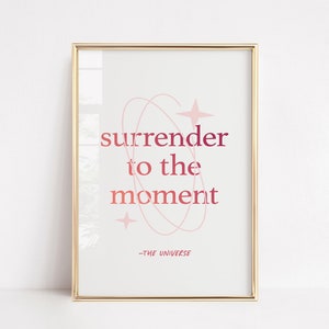 Puede incluir: Una impresión enmarcada en dorado con fondo blanco y texto rosa y rojo que dice "surrender to the moment -THE UNIVERSE". El texto está rodeado por un diseño abstracto rosa y rojo.