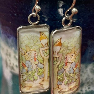 Pode incluir: Um par de brincos de prata com pingentes retangulares de vidro que apresentam uma ilustração do Peter Rabbit. Os pingentes representam o Peter Rabbit segurando uma cenoura e um pássaro empoleirado em um poste de cerca.