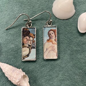 Könnte beinhalten: Ein Paar silberne Ohrringe mit rechteckigen Glasplatten, die Bilder der Renaissancekunst zeigen. Der linke Ohrring zeigt eine Frau mit langen braunen Haaren und einem blauen Kleid, während der rechte Ohrring eine Frau mit langen blonden Haaren und einem nackten Körper zeigt.