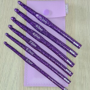 Op de afbeelding: Set van zes paarse haaknaalden met glitter, variërend in grootte van 5,0 mm tot 10,0 mm. De haaknaalden zitten in een paarse etui met de merknaam "emce" erop gedrukt.
