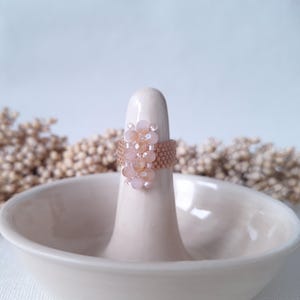 Op de afbeelding: Een delicate ring met een cluster van lichtroze en perzikkleurige kralen, geplaatst op een witte keramische ringhouder.