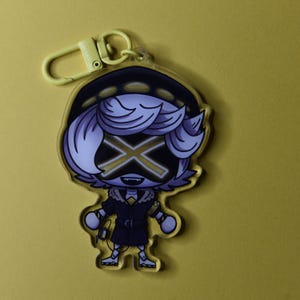 May include: A yellow-toned keychain featuring a cartoon character with a black and gold X-shaped face. The character wears a dark coat with a fur collar and a hat. The keychain has a yellow clasp.