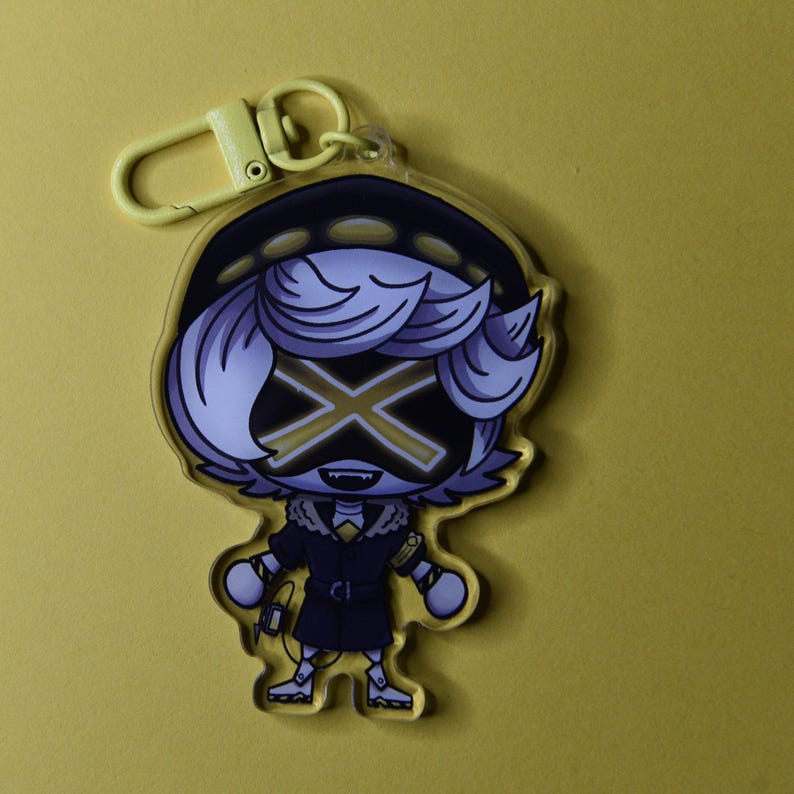 May include: A yellow-toned keychain featuring a cartoon character with a black and gold X-shaped face. The character wears a dark coat with a fur collar and a hat. The keychain has a yellow clasp.