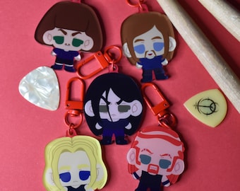 May include: Five cartoon character keychains with red clasps, each featuring a different character design. Two guitar picks and two drumsticks are also visible. The keychains are on a red background.