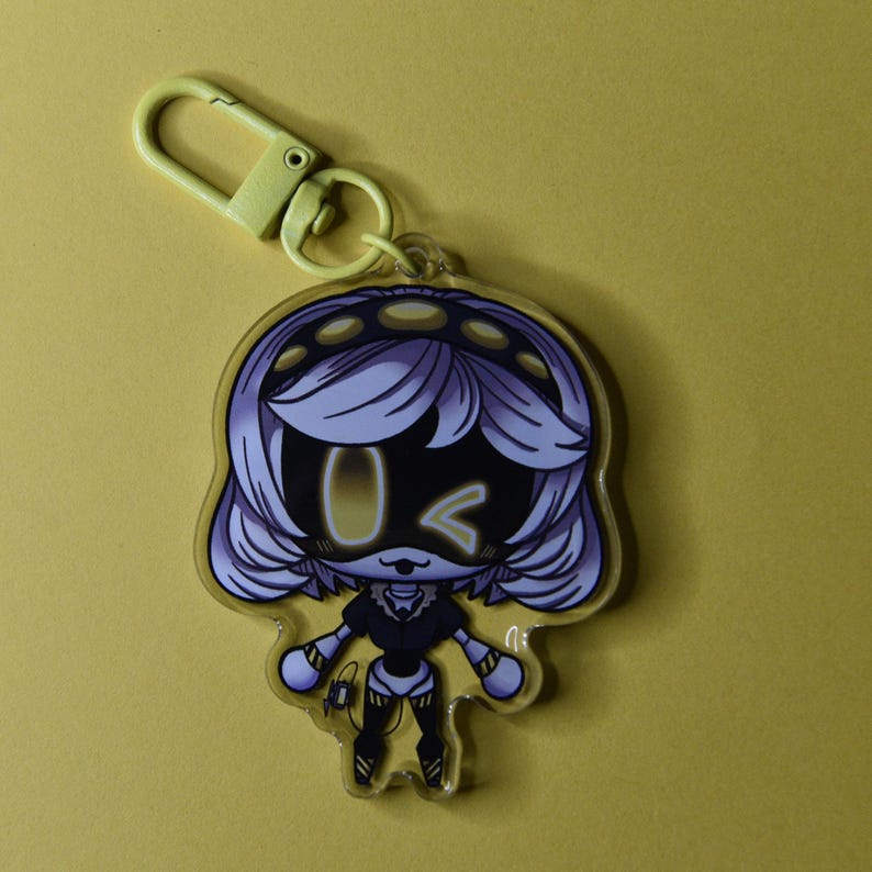 May include: A cartoon-style keychain featuring a character with white hair, a black face, and yellow accents. The keychain has a yellow clasp and a yellow background. The character is in a standing pose with a winking expression.