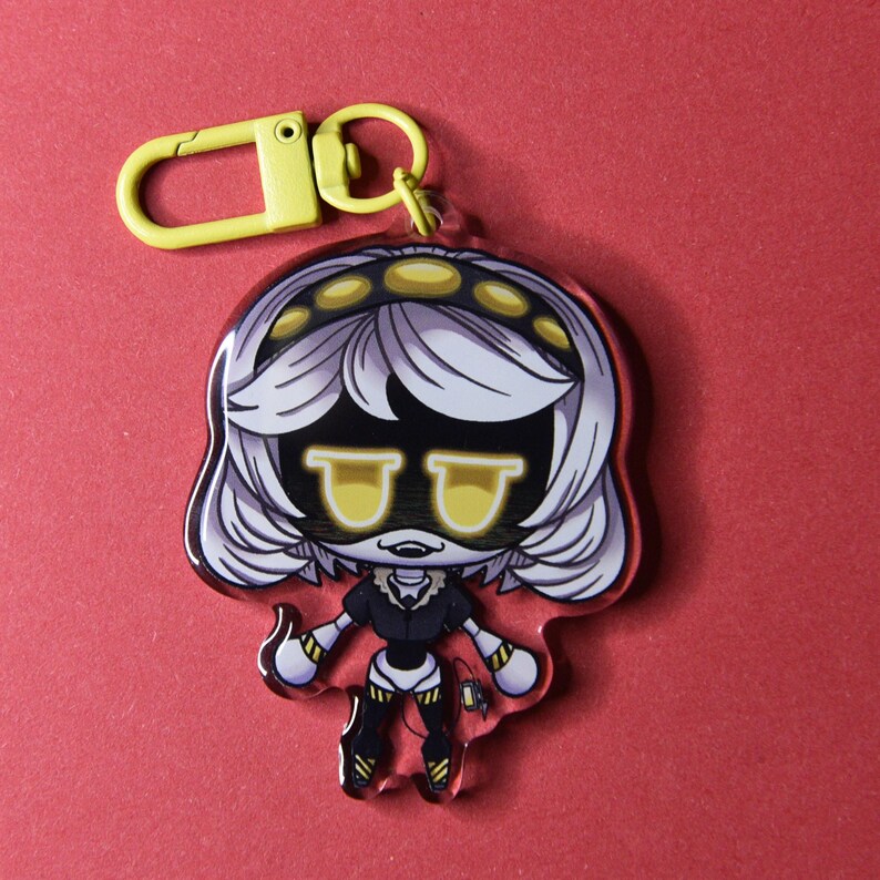 May include: A clear acrylic keychain featuring a cartoon character with white hair, yellow goggles, and a black outfit. The keychain has a yellow clasp and ring. The character is set against a red background.