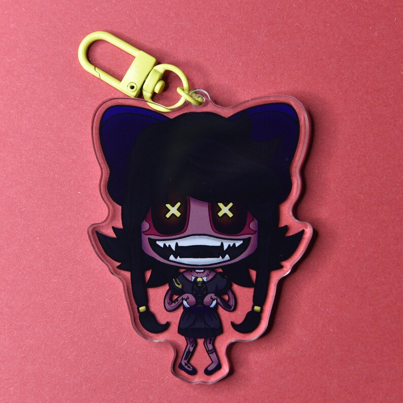 May include: A clear acrylic keychain featuring a cartoon character with black hair, a dark dress, and yellow eyes. The character has a wide, toothy grin and is attached to a yellow metal clasp. The background is a solid red.
