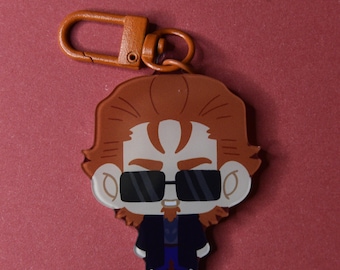 May include: A novelty keychain featuring a cartoon character with sunglasses, a beard, and brown hair. The keychain has a brown clasp and a red background.