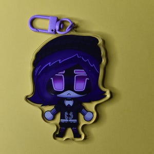 May include: A clear acrylic keychain featuring a cartoon character with purple hair, a black hat, and a black outfit. The character has purple eyes and a stern expression. A purple clasp is attached to the top.