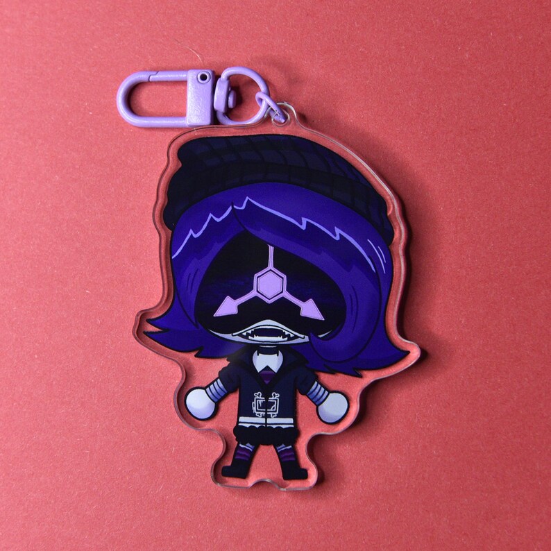 May include: A clear acrylic keychain featuring a cartoon character with purple hair, a black hat, and a black outfit. The character has a white face with a geometric design and a purple clasp. The background is a solid red.