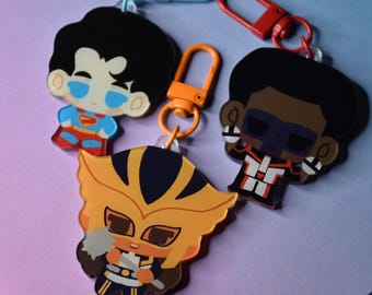 May include: Three cartoon character keychains with colorful clasps. One keychain features a character in a blue and red suit, another in a black and white suit, and the third in a yellow and brown costume. The clasps are blue, red, and orange.