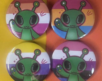 May include: Four round buttons featuring a green cartoon alien with large eyes and antennae. Each button has a different background color scheme, including pink, orange, yellow, purple, and white, representing various pride flags. The alien is waving.