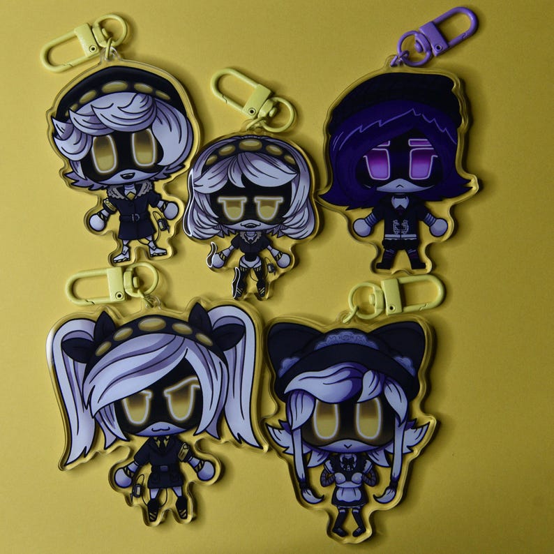 May include: Five acrylic keychains featuring cartoon characters with yellow eyes and accents. Each keychain has a different character design, with various hairstyles and outfits. The keychains have gold and purple clasps, and are set against a yellow background.