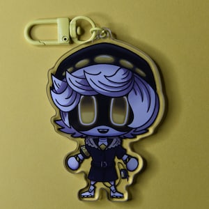 May include: A yellow keychain featuring a cartoon character with light purple hair, a black outfit, and yellow eyes. The character has a black hat and a yellow clasp. The background is a solid yellow color.