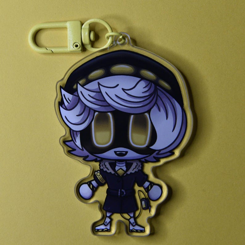 May include: A yellow keychain featuring a cartoon character with light purple hair, a black outfit, and yellow eyes. The character has a black hat and a yellow clasp. The background is a solid yellow color.