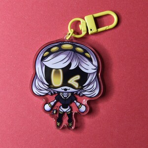 May include: A clear acrylic keychain featuring a cartoon character with white hair, a black and yellow outfit, and yellow accents. The keychain has a yellow clasp and a red border. The background is a solid red color.