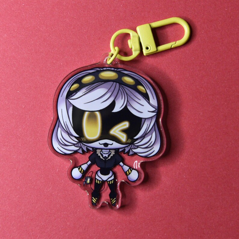 May include: A clear acrylic keychain featuring a cartoon character with white hair, a black and yellow outfit, and yellow accents. The keychain has a yellow clasp and a red border. The background is a solid red color.