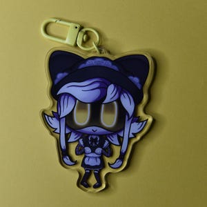 May include: A clear acrylic keychain featuring a cartoon character with a black hat, dress, and yellow eyes. The character has white hair and wings. The keychain has a yellow clasp.
