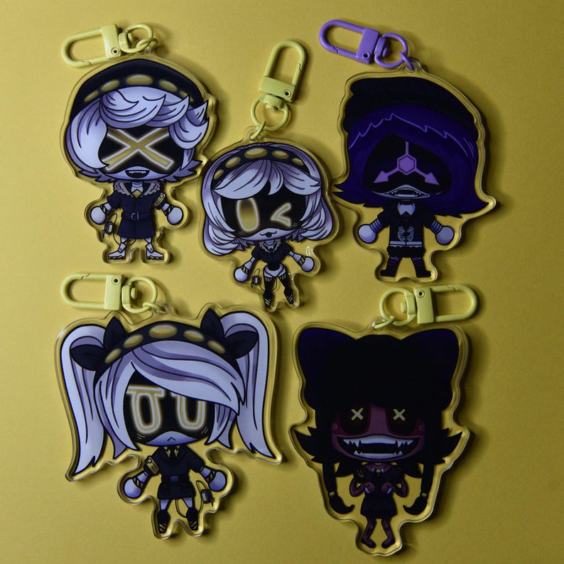 May include: A collection of five acrylic keychains featuring cartoon characters with yellow and black accents. Each keychain has a different character design, with various hairstyles and facial expressions. The keychains are attached to yellow and purple clasps.