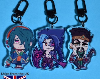 May include: Three acrylic keychains featuring cartoon characters with black carabiner clips. The characters have detailed designs with vibrant colors. The background is a solid light blue. The text "Ships from the UK" is visible.