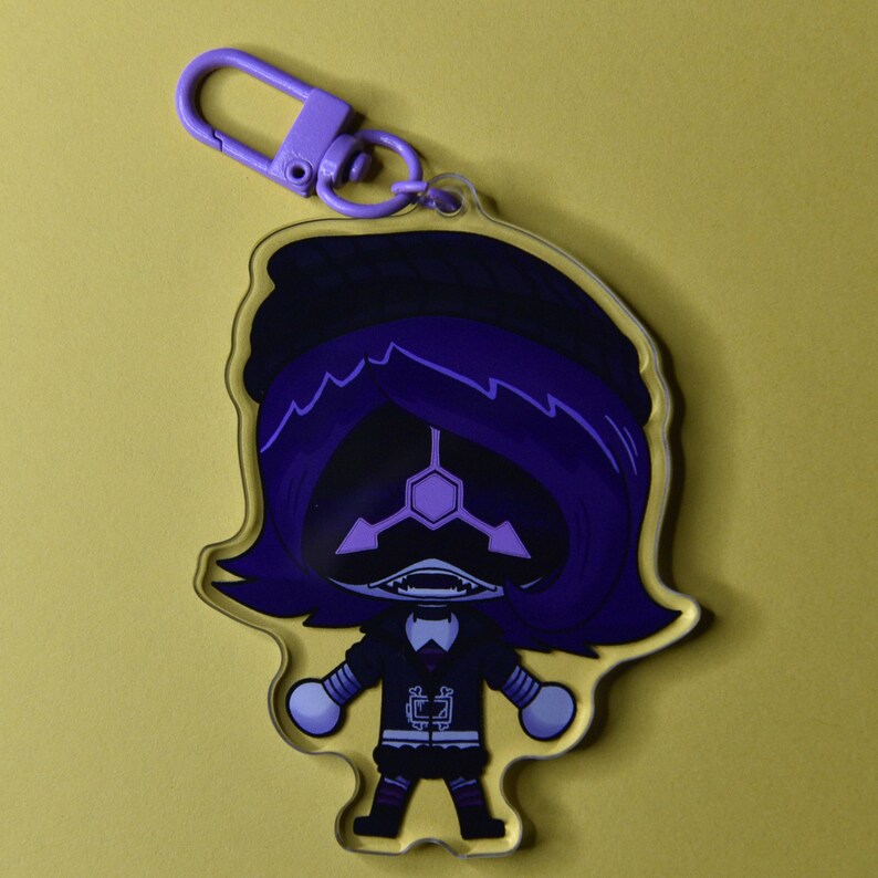 May include: A clear acrylic keychain featuring a cartoon character with purple hair and a black outfit. The character has a geometric face design and is attached to a purple clasp.