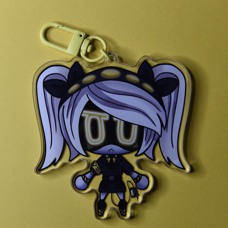 May include: A clear acrylic keychain featuring a cartoon character with white hair, black accents, and yellow highlights. The character has a black outfit and a yellow clasp. The background is a solid yellow.