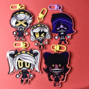 May include: A collection of five acrylic keychains featuring cartoon characters with white hair, black outfits, and yellow accents. Each keychain has a different character design and is attached to a yellow or purple clasp.