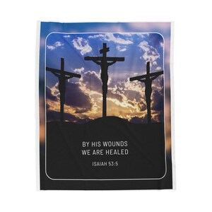 Puede incluir: Una manta de tiro con temática religiosa con una silueta de tres cruces contra un cielo de puesta de sol nublado. El texto "By His Wounds We Are Healed" e "Isaiah 53:5" está impreso debajo de las cruces.