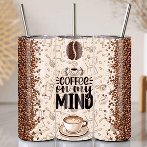 May include: A stainless steel travel mug with a coffee-themed design. The mug features a repeating pattern of coffee beans and coffee-making equipment. The phrase "coffee on my MIND" is displayed. The mug includes a silver straw.