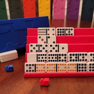 May include: A red and blue domino rack with a train design. The rack is filled with white dominoes with black dots. There are two small toy trains on the table.