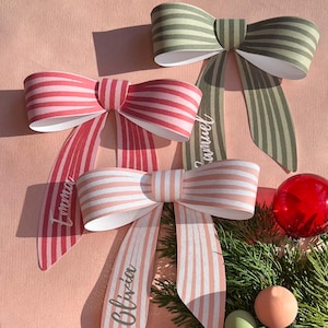 Peut inclure: Trois nœuds décoratifs en papier à rayures roses, vertes et pêche. Chaque nœud a de longs rubans avec des noms imprimés. Une boule rouge et de la verdure sont en arrière-plan.