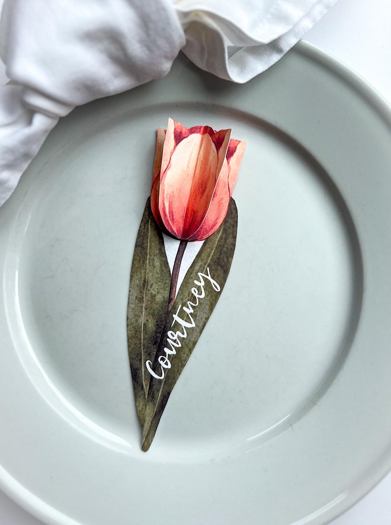 May include: A paper tulip place card with a green stem and leaves. The name "Courtney" is written in white script on the leaf.