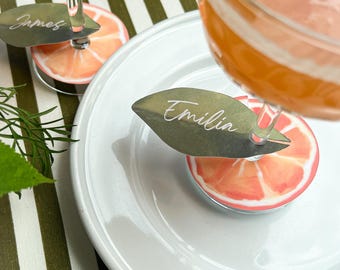 May include: Two clear stemmed cocktail glasses filled with an orange liquid, each featuring a circular citrus slice base and a green leaf-shaped name tag attached to the stem. The tags display the names James and Emilia in white script lettering.