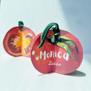 May include: Two tomato-shaped paper ornaments with green ribbon hangers. One is open, revealing the interior. The other features the name "Monica Russa" in white script on a red background with green leaves.
