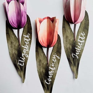 May include: Three paper tulip place cards with watercolour designs. The tulips are pink, orange, and purple. The names "Elizabeth", "Courtney", and "Juliette" are written on the stems.