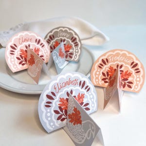 May include: Decorative place cards with names such as "Claire" and "Elizabeth" printed on them. The cards have a half-circle design with autumnal leaf and floral patterns in orange, pink, and brown. Each card has a folded base for standing.