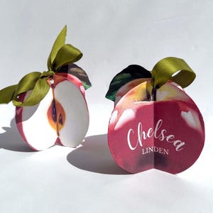 Op de afbeelding: Twee rode, appelvormige geschenkdozen met groene strikken. Op één doos staat de naam "Chelsea Linden".
