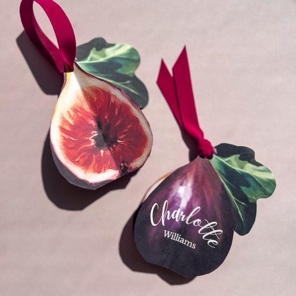May include: Two fig-shaped gift tags with a deep red ribbon. One tag shows a fig's cross-section, revealing the red interior. The other is a dark purple fig with the name "Charlotte Williams" in white script.