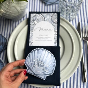 May include: A table setting with a navy blue napkin, white plates, and a menu card. A seashell-shaped place card with the name "Rebecca Miller" is held above the napkin. The background features a blue and white striped tablecloth.