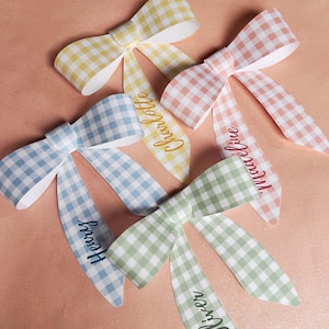May include: Four pastel-coloured gingham bows in yellow, pink, blue, and green. Each bow has a name printed on one of the tails: Charlotte, Madeline, Henry, and Oliver. The bows are arranged on a light pink surface.