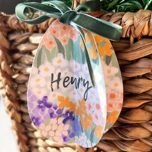 Könnte beinhalten: Ein geflochtener Korb mit einem grünen Samtband, das um ein blumenförmiges Etikett gebunden ist. Das eiförmige Etikett trägt den Namen "Henry" in schwarzer Schrift. Das florale Design umfasst orange, lila und weiße Blumen.