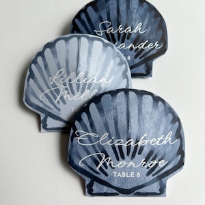 May include: Three seashell-shaped place cards with watercolor-style blue and gray designs. Each card features a guest's name in elegant white script, along with "TABLE 8". Ideal for a beach-themed event.