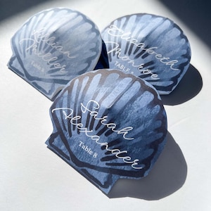 May include: Three seashell-shaped place cards with watercolor blue and gray designs. Each card features a different name in elegant white script: Lillian Miller, Elizabeth Monroe, and Sarah Alexander. The text "Table 8" is also included.