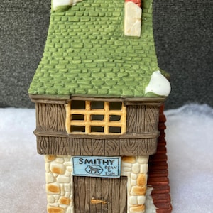 以下が含まれることがあります： 緑色の屋根と煙突のある、小さく、茶色と白のセラミック製の建物。建物には「Smithy Bean & Son」と書かれた看板があります。