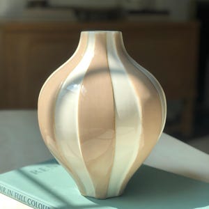 May include: A decorative ceramic vase with vertical stripes in alternating shades of beige and white. The vase has a bulbous shape and a narrow neck. It is sitting on a book with the text "IN FULL COLOUR - PHAIDON".